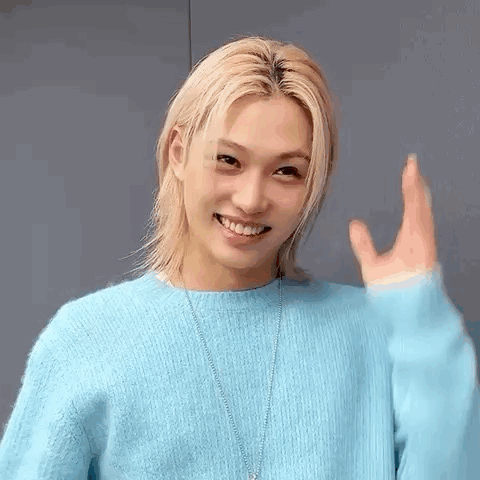 A Young Man With Long Blonde Hair Is Wearing A Blue Sweater And Necklace . GIF