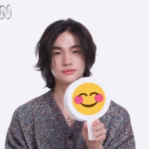 A Young Man With Long Hair Is Holding A Smiley Face With Pink Cheeks . GIF