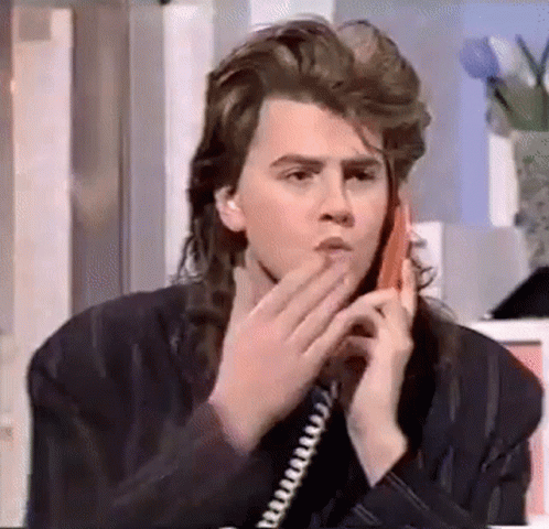 A Young Man With Long Hair Is Talking On A Telephone . GIF