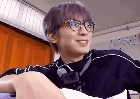 A Young Man With Purple Hair And Glasses Is Sitting On A Couch . GIF
