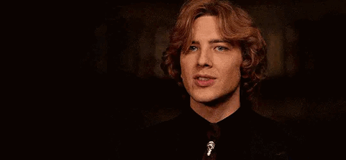 A Young Man With Red Hair Is Wearing A Black Shirt And Tie And Looking At The Camera . GIF