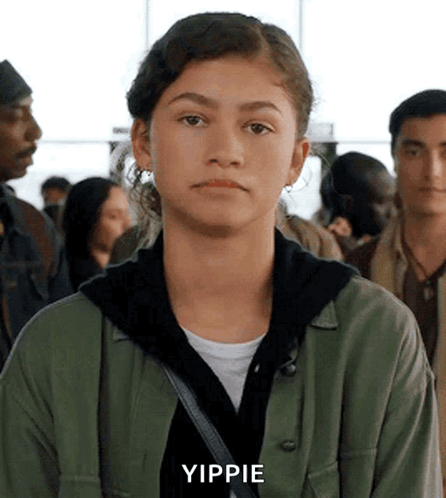A Young Woman In A Green Jacket Is Standing In A Crowd Of People And Making A Funny Face . GIF