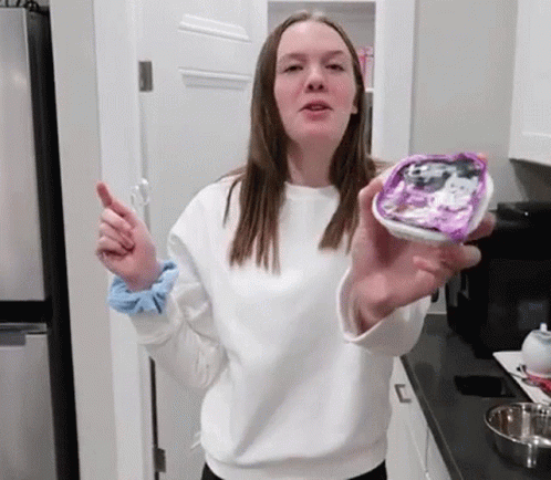 A Young Woman In A White Sweater Is Holding A Container Of Food In Her Hand . GIF