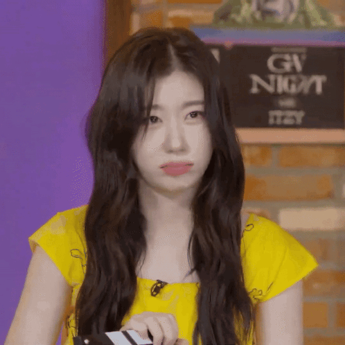A Young Woman In A Yellow Shirt Is Sitting In Front Of A Sign That Says Gw Night GIF