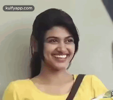 A Young Woman In A Yellow Shirt Is Smiling And Looking At The Camera . GIF