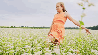 A Young Woman In An Orange Dress Is Running Through A Field Of White Flowers . GIF