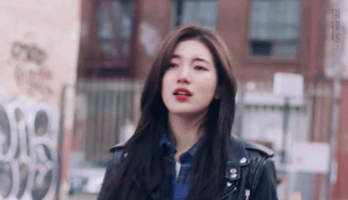 A Young Woman Wearing A Leather Jacket And A Plaid Shirt Is Standing In Front Of A Building . GIF