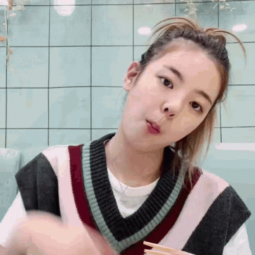 A Young Woman Wearing A Sweater Vest And A Ponytail Is Eating With Chopsticks . GIF