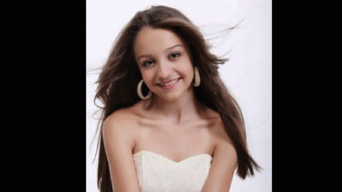 A Young Woman Wearing A White Strapless Dress And Hoop Earrings Smiles GIF