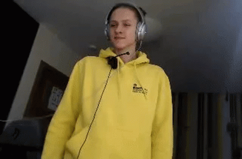 A Young Woman Wearing A Yellow Hoodie And Headphones Is Standing In A Room . GIF