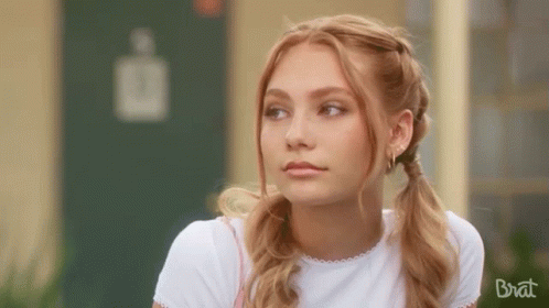 A Young Woman With Pigtails And Earrings Is Sitting In Front Of A Building . GIF
