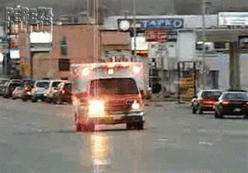 An Ambulance Is Driving Down A Street In Front Of A Tapco GIF