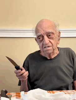 An Elderly Man Is Sitting At A Table Holding A Knife In His Hand GIF