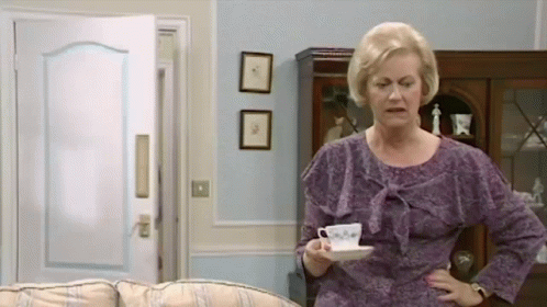 An Elderly Woman In A Purple Dress Is Holding A Cup Of Coffee In A Living Room . GIF