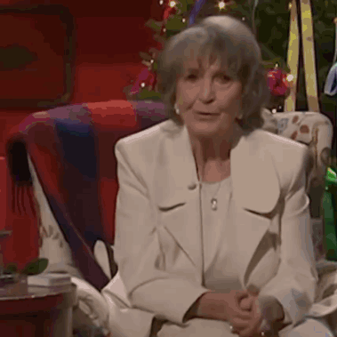 An Elderly Woman In A White Suit Sits In A Chair In Front Of A Christmas Tree GIF