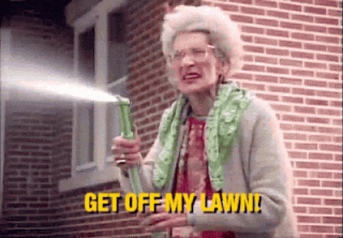 An Elderly Woman Is Holding A Hose In Front Of A Brick Building And Saying `` Get Off My Lawn '' . GIF