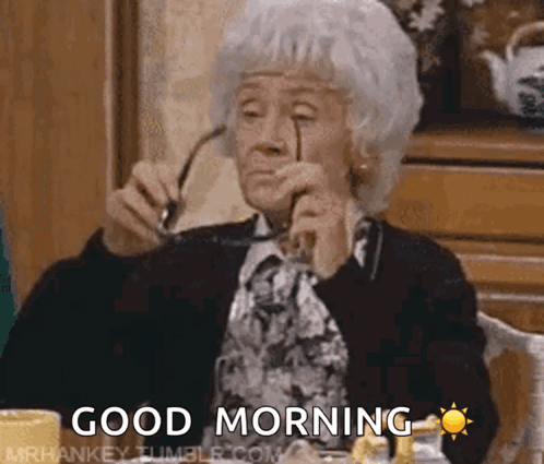 An Elderly Woman Is Sitting At A Table Holding Glasses And Says Good Morning GIF