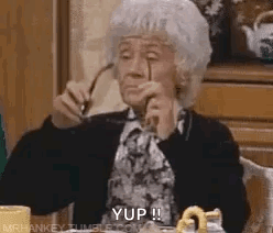 An Elderly Woman Is Sitting At A Table Holding Glasses In Her Hand . GIF