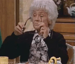 An Elderly Woman Is Sitting At A Table Holding Glasses In Her Hand . GIF
