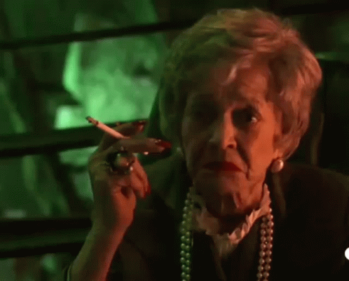 An Elderly Woman Is Smoking A Cigarette And Waving At The Camera In Front Of A Green Background . GIF
