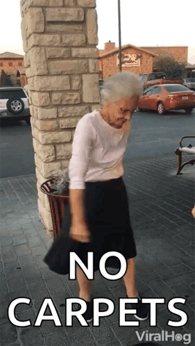 An Elderly Woman Is Standing In Front Of A Brick Wall With The Words No Carpets Written On It GIF