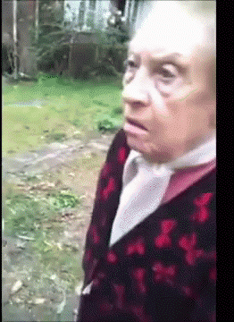 An Elderly Woman Wearing A Red And Black Sweater Is Standing In A Field GIF