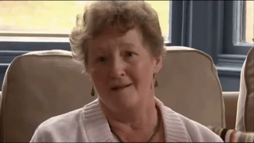An Elderly Woman Wearing A White Sweater And Earrings Is Sitting On A Couch GIF