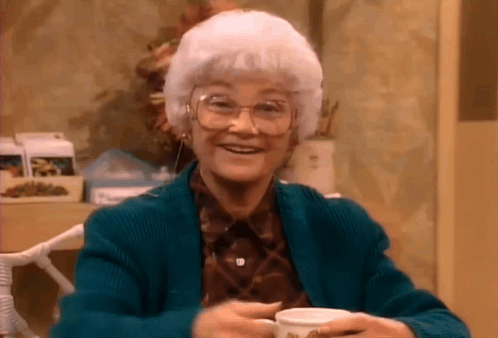 An Elderly Woman Wearing Glasses And A Blue Sweater Is Holding A Cup Of Coffee And Smiling . GIF