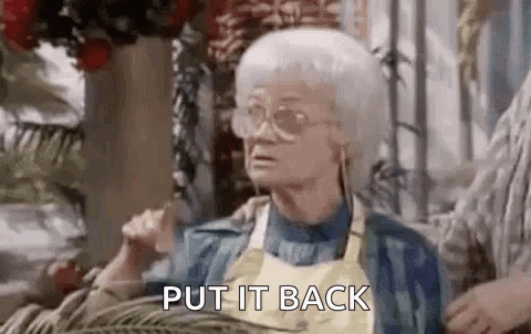 An Elderly Woman Wearing Glasses And A Yellow Apron Is Sitting On A Couch And Saying `` Put It Back '' . GIF