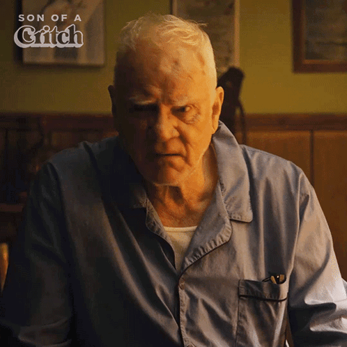 An Older Man In A Blue Shirt Is Sitting In Front Of A Sign That Says Son Of A Grifch GIF