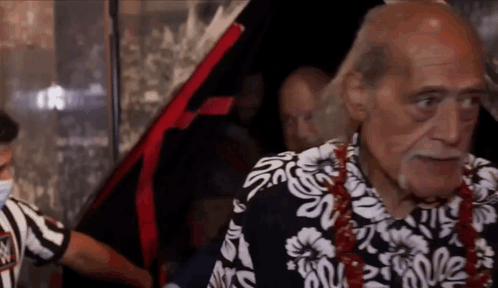 an older man wearing a hawaiian shirt is standing in front of a car .