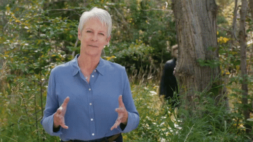 An Older Woman In A Blue Shirt Is Standing In The Woods GIF
