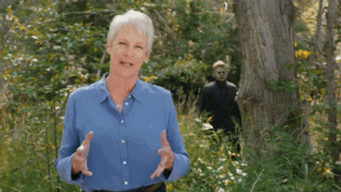 An Older Woman In A Blue Shirt Is Standing In The Woods Talking To Someone . GIF