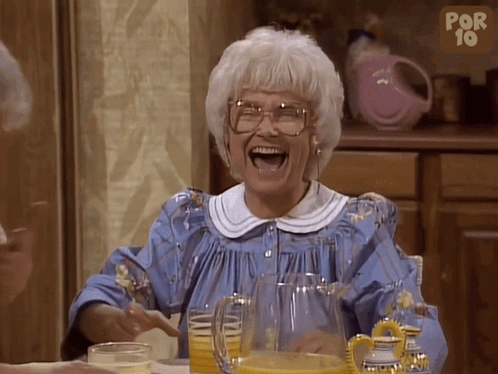 An Older Woman Is Laughing With A Pitcher Of Orange Juice In Front Of Her GIF