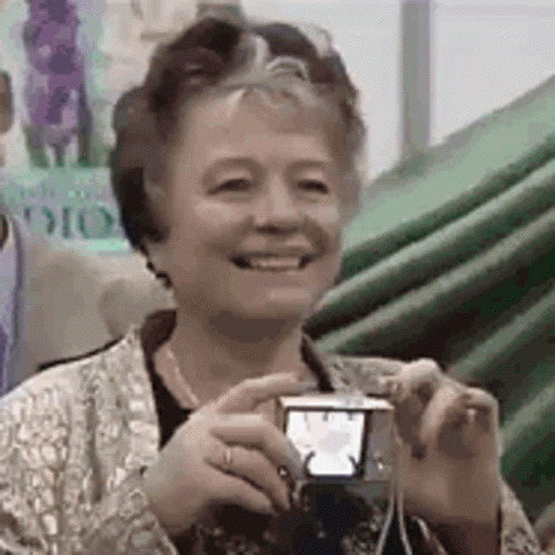 An Older Woman Is Smiling While Holding A Small Camera . GIF