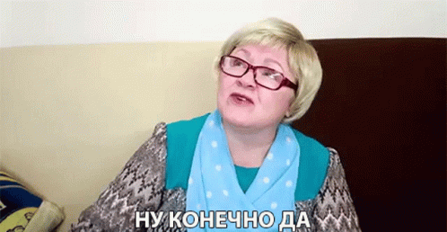 An Older Woman Wearing Glasses And A Blue Scarf Is Sitting On A Couch And Talking In Russian . GIF