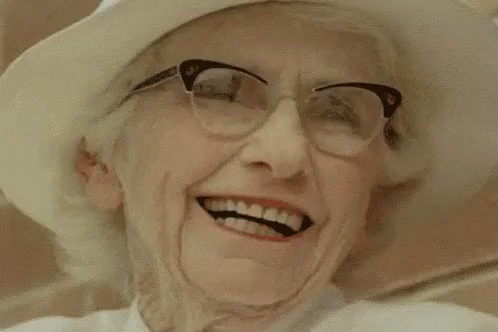 An Older Woman Wearing Glasses And A White Hat Is Smiling . GIF