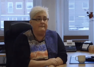 An Older Woman Wearing Glasses Sits At A Desk With Her Hands Folded GIF