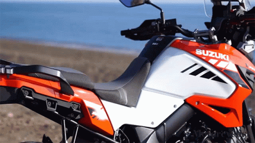 An Orange And White Suzuki Motorcycle Is Parked On A Beach GIF