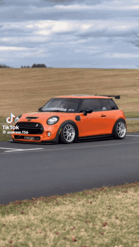 An Orange Mini Cooper Is Parked On The Side Of The Road With A Field In The Background GIF