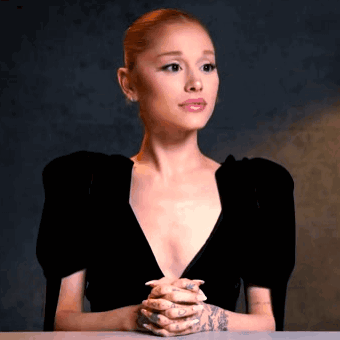 Ariana Grande Is Sitting At A Table With Her Hands Folded In Front Of Her Face . GIF