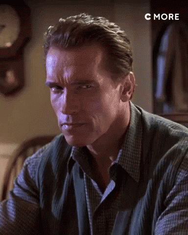Arnold Schwarzenegger Is Sitting At A Table In Front Of A Clock And Looking At The Camera . GIF