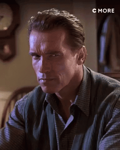 Arnold Schwarzenegger Is Sitting At A Table In Front Of A Clock And Looking At The Camera . GIF