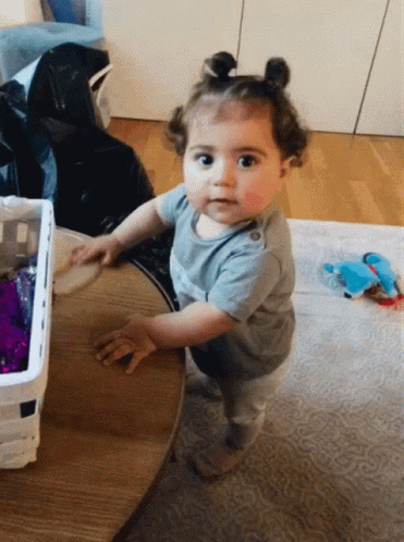 Baby Girl Standing at Wooden Table Looking at Camera