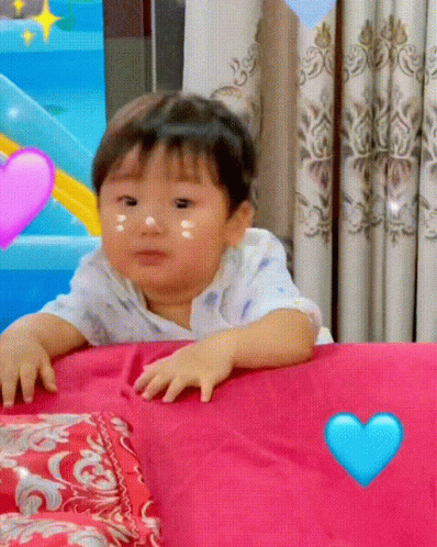 Baby Sitting On Bed With Pink Pillow And Blue Heart GIF