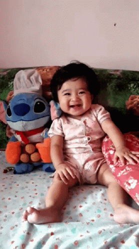 Baby Sitting On Bed With Stitch Stuffed Animal GIF