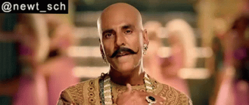 Bald Man In Gold Dress With Mustache, Earrings, And Necklace GIF