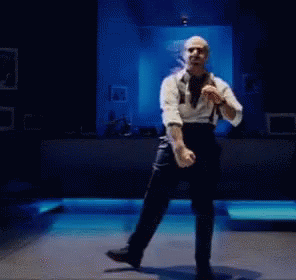Bald Man In White Shirt And Black Pants Dancing On Blue Stage GIF