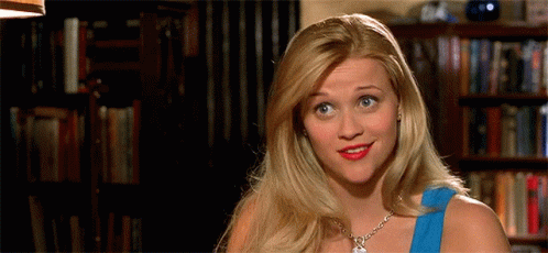 Blonde Woman In Blue Top Smiling In Front Of Bookshelf GIF