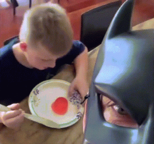 Boy Eating Plate of Food Next to Batman Mask